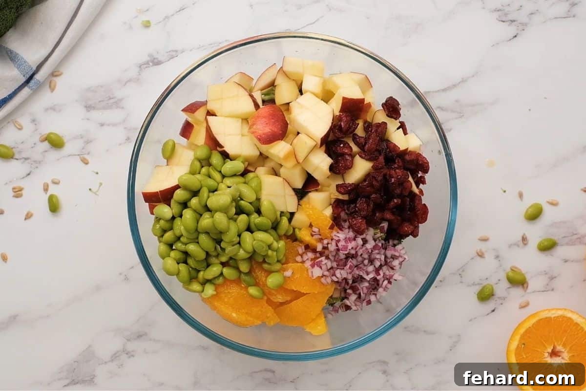 A large mixing bowl filled with the colorful raw ingredients for the broccoli apple salad, including chopped broccoli, diced apples, orange segments, edamame, red onion, and cranberries, before the dressing is added.