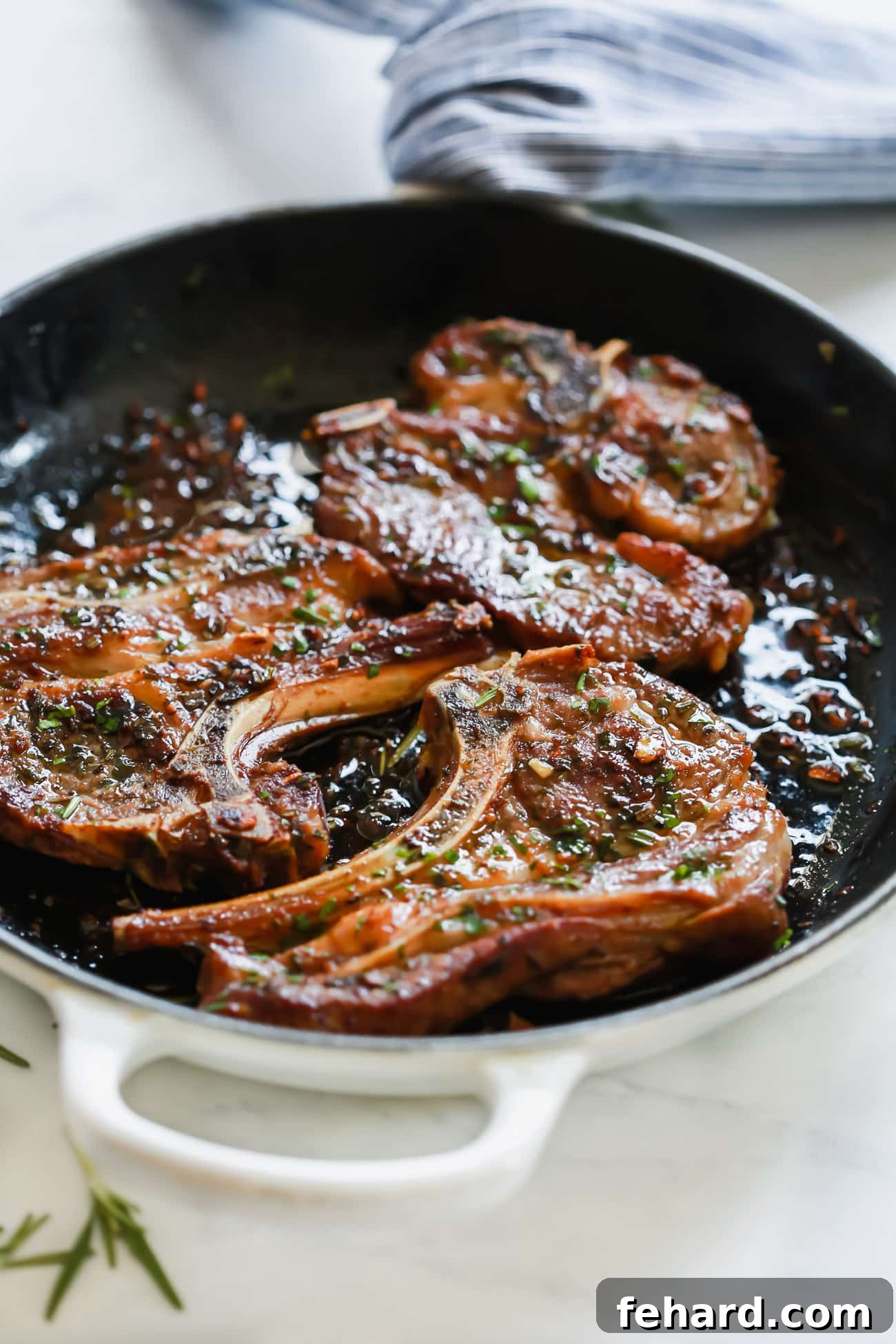 A close up of a white cast iron skillet containing perfectly seared Garlic Herb Lamb Chops, garnished with fresh herbs.