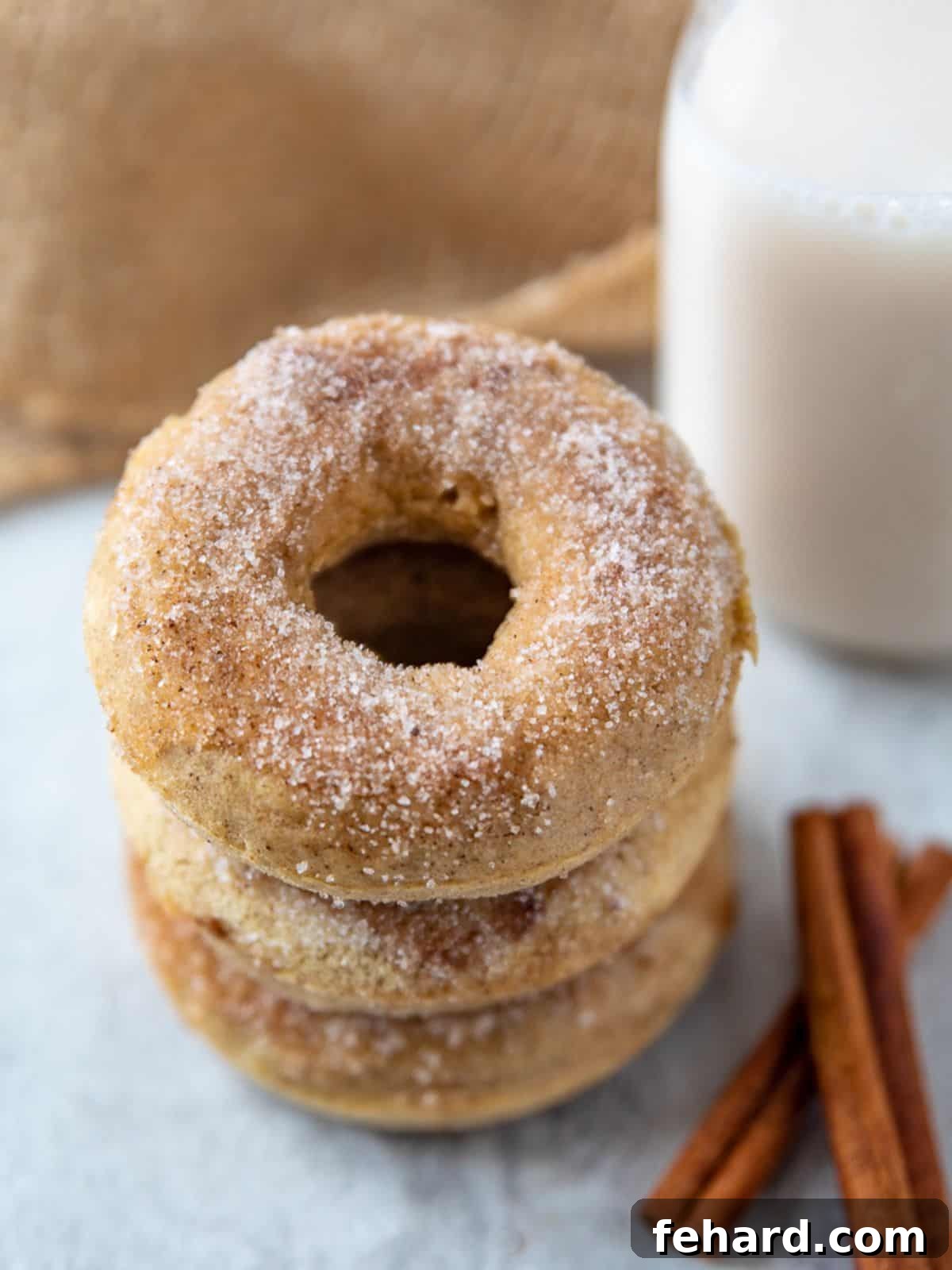 Three golden-brown baked donuts generously coated in sparkling cinnamon sugar, stacked elegantly beside rustic cinnamon sticks and a tall glass of milk, inviting a delightful breakfast or snack.