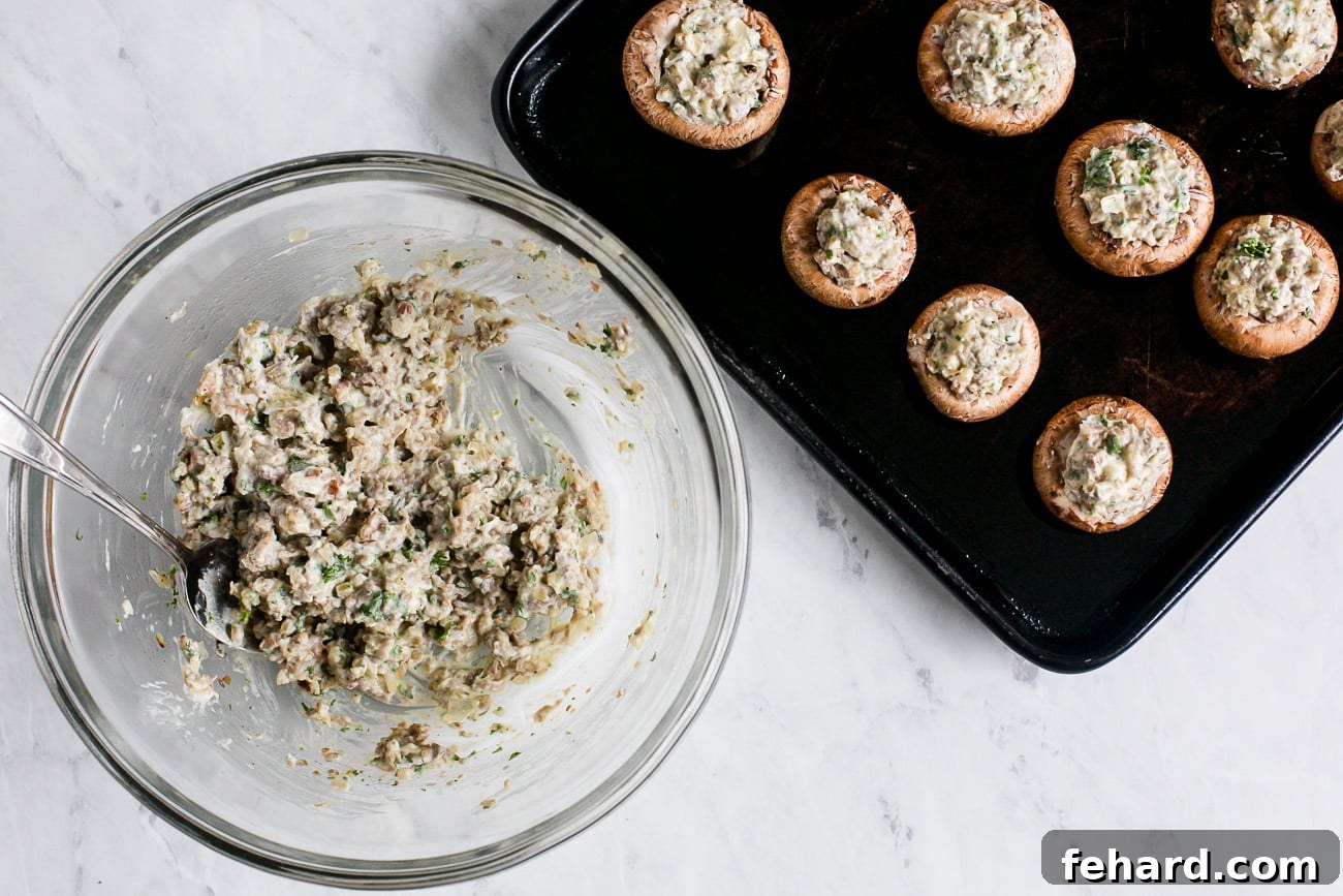 A baking sheet filled with hollowed mushrooms being stuffed with the creamy sausage filling.
