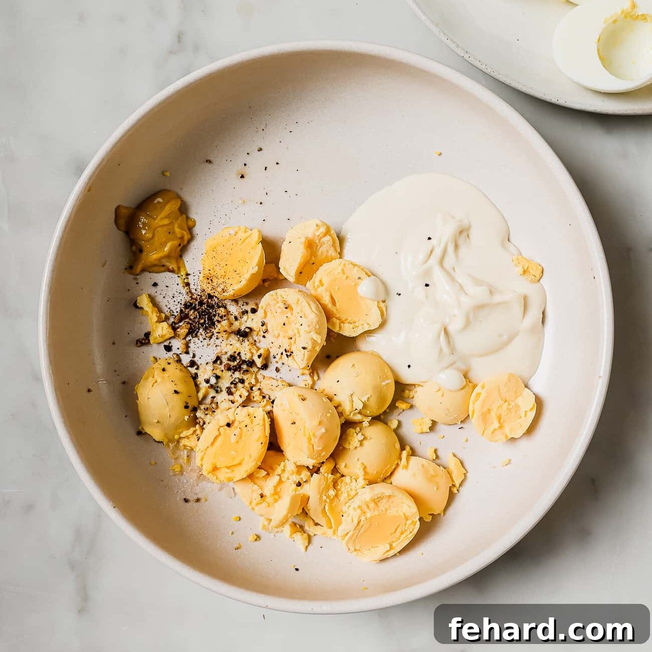 A bowl containing scooped egg yolks, surrounded by small dishes of mayonnaise, apple cider vinegar, salt, pepper, and garlic powder, ready to be mixed for the deviled egg filling.