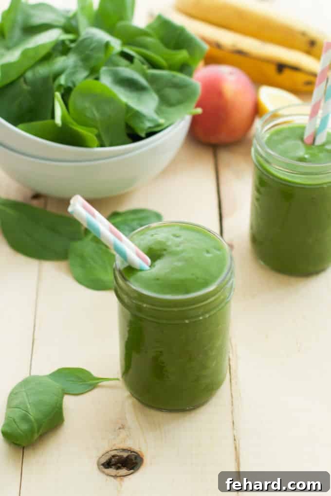 Two glasses of peach green smoothie beside an apple, bananas, and a bowl of spinach, showcasing fresh ingredients.