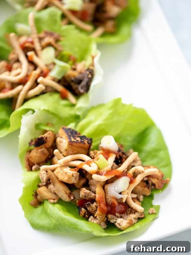 A vibrant lettuce cup filled with a golden, crispy tofu and mushroom vegetarian filling, generously topped with crunchy chow mein noodles and a drizzle of hot sauce, ready to be enjoyed.