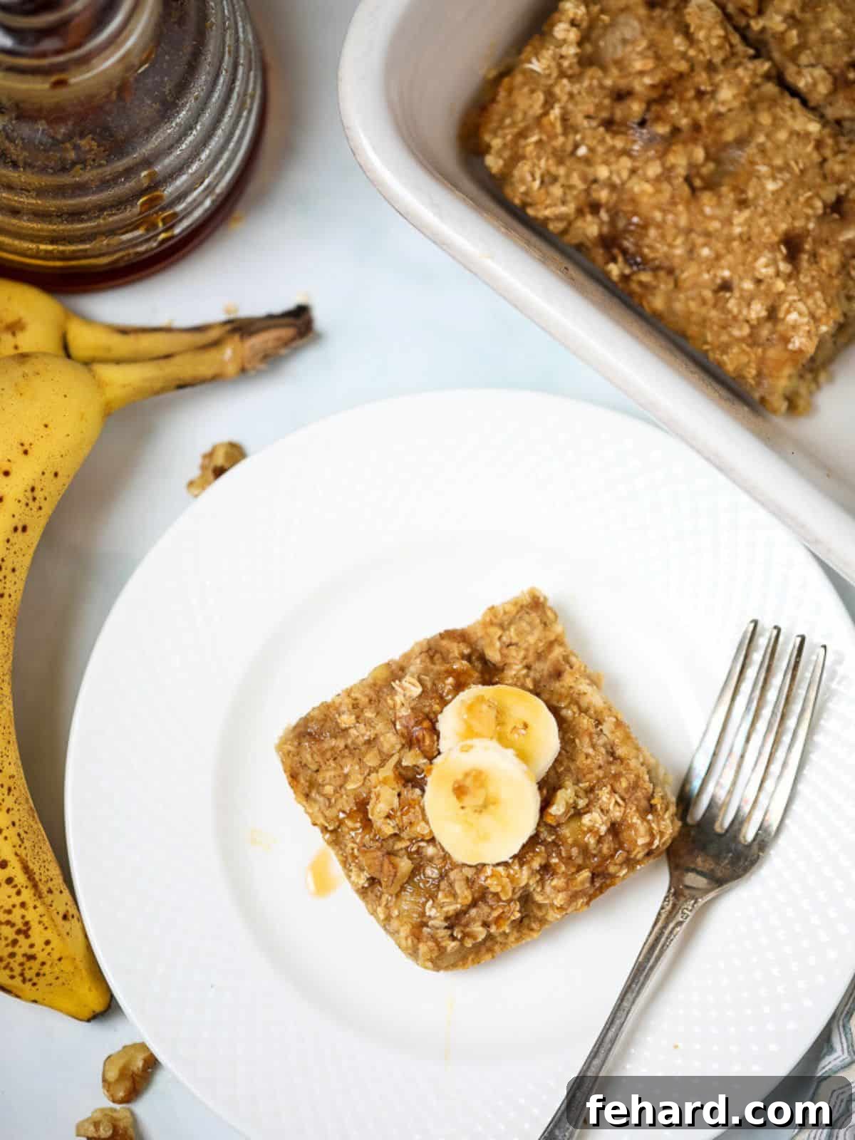 A golden brown slice of banana baked oatmeal sits on a pristine white plate, elegantly garnished with fresh banana slices and a delicate drizzle of honey. Ripe bananas rest invitingly beside it, hinting at the key ingredient.