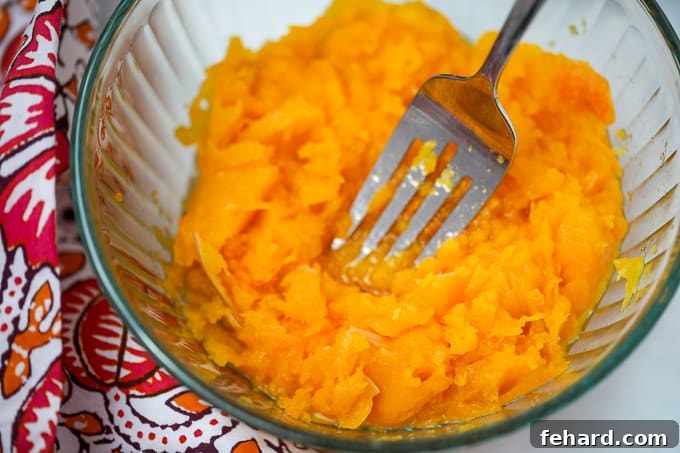 Butternut squash being mashed with a fork in a clear glass bowl, showcasing its soft texture.