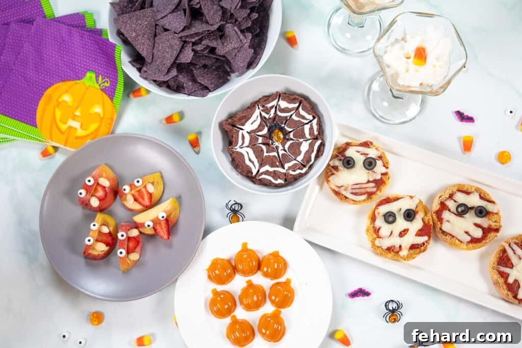 A vibrant display of various healthy Halloween snacks, including apple monsters, mummy pizzas, and a spiderweb dip, arranged on a white counter with festive Halloween decorations.