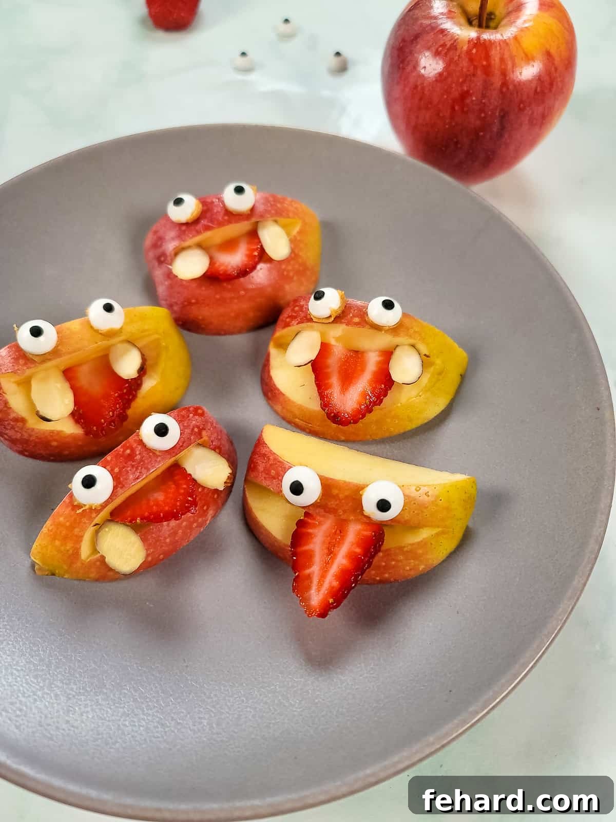 Cute apple slices decorated to look like friendly monsters with strawberry tongues and candy eyes, arranged on a gray plate.