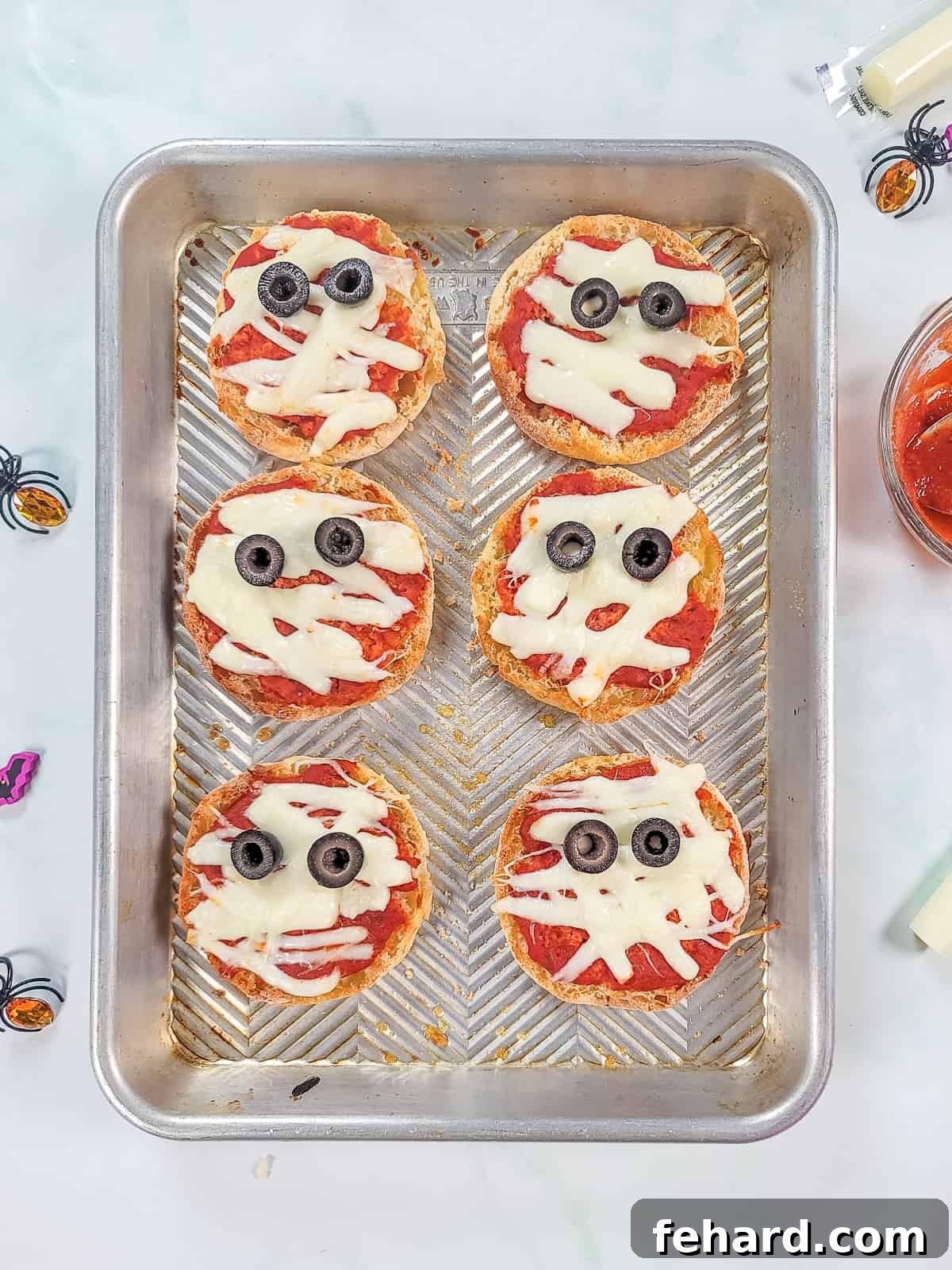 A baking sheet filled with individual English muffin pizzas designed to look like mummies with cheese strips and olive eyes.