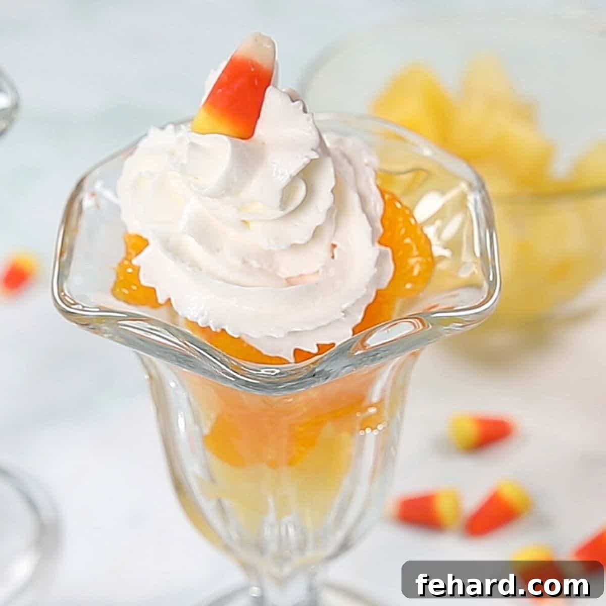 A clear glass parfait dish showcasing distinct layers of pineapple, mandarin oranges, and whipped cream, topped with a piece of candy corn.