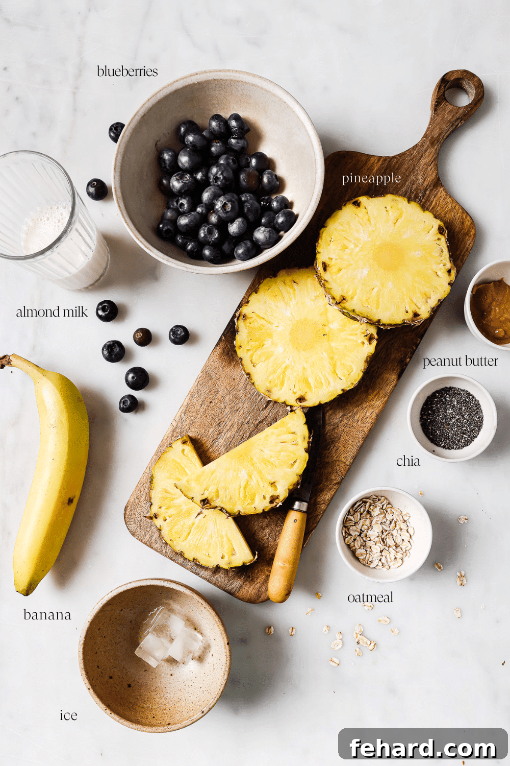 Pre-measured ingredients for a blueberry banana smoothie arranged on a white countertop, including frozen banana, fresh blueberries, pineapple, almond milk, peanut butter, chia seeds, and oats.