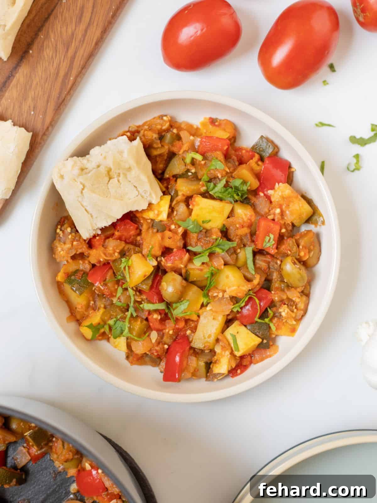 Bowl of thick homemade ratatouille in white bowl with rustic piece of bread, garnished with fresh basil.