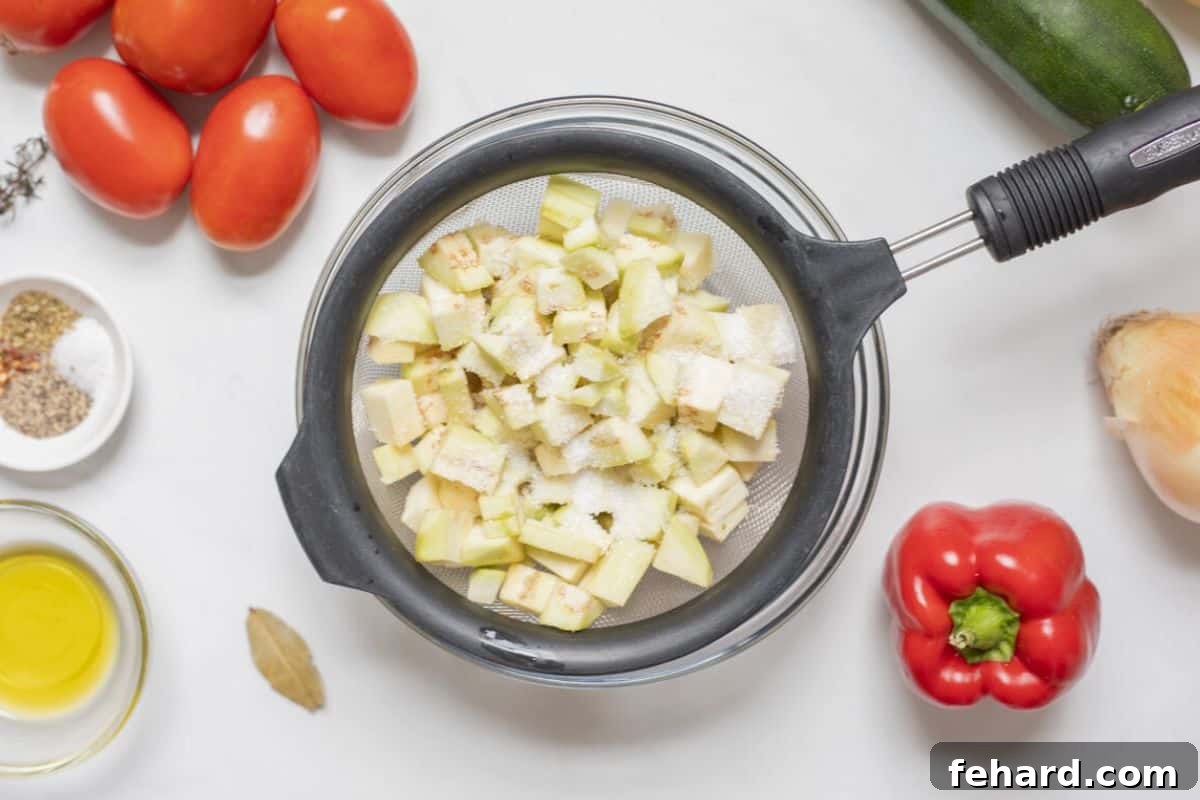 Peeled and cubed eggplant in a fine mesh strainer, next to other ingredients for Classic Ratatouille.
