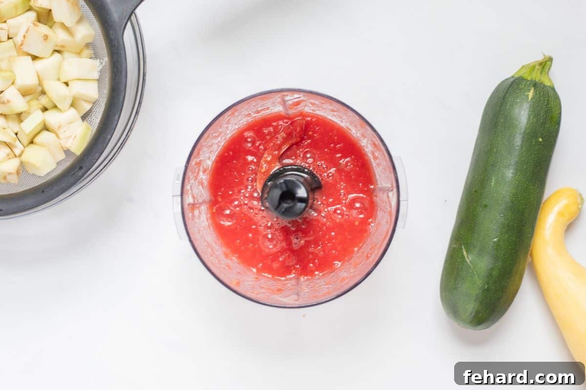 Pureed fresh tomatoes in a small food processor, next to sliced summer squash.