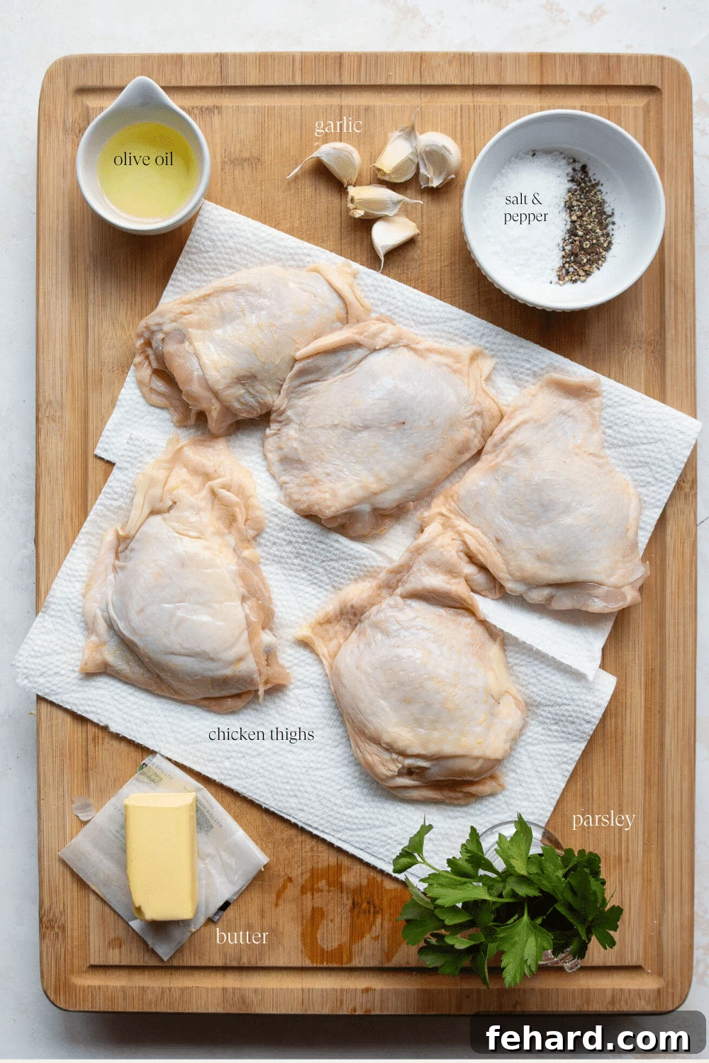 Overhead view of ingredients on a wooden board: chicken thighs, garlic cloves, butter, salt, pepper, and olive oil.