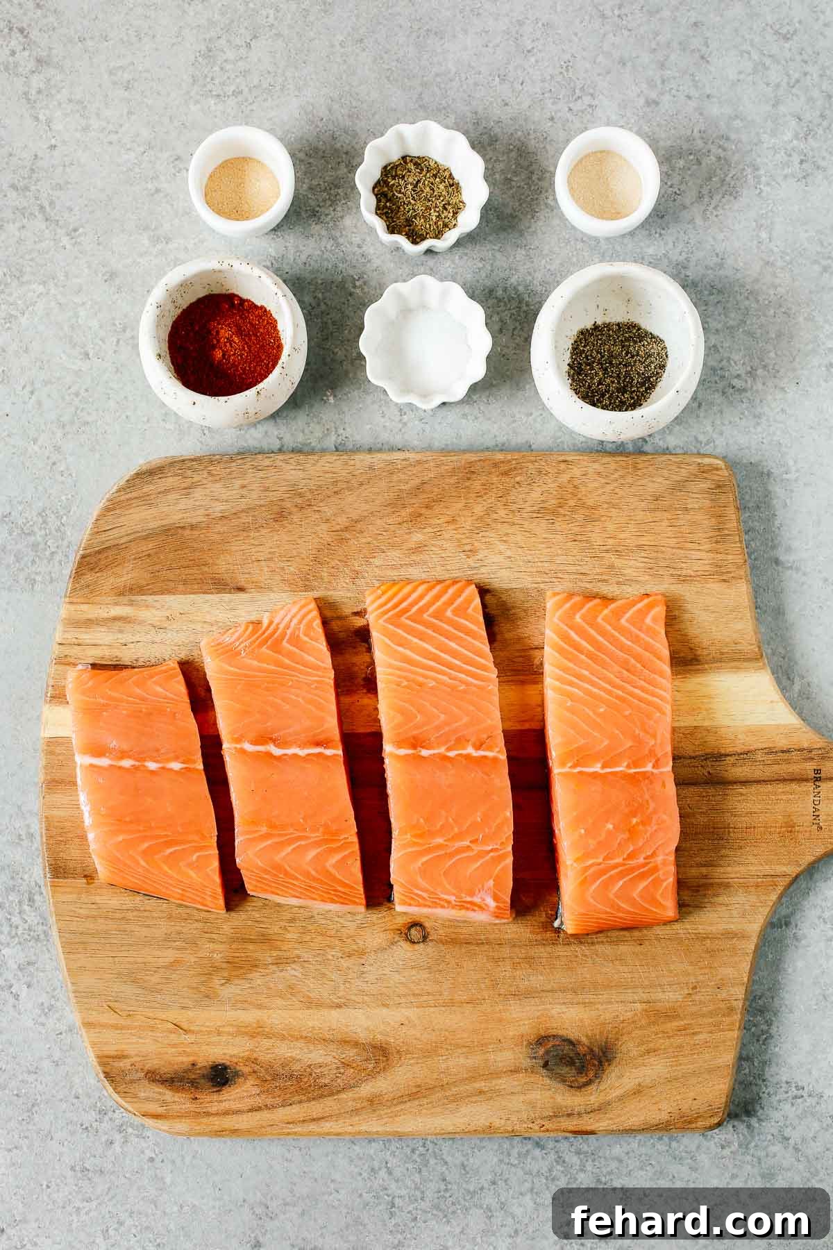 Overhead view of four raw salmon fillets laid out on a cutting board, surrounded by six small bowls filled with various spices, ready to be combined for the blackening seasoning blend.