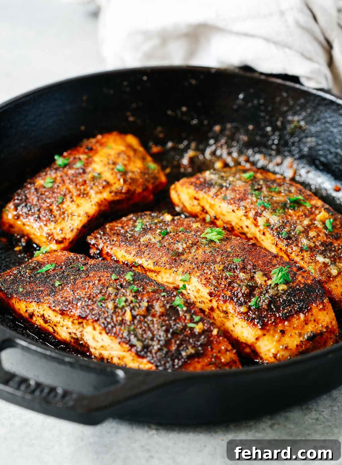 Close-up view of four heavily seasoned salmon fillets sizzling and cooking in a hot cast iron skillet, with visible steam and crisping edges.