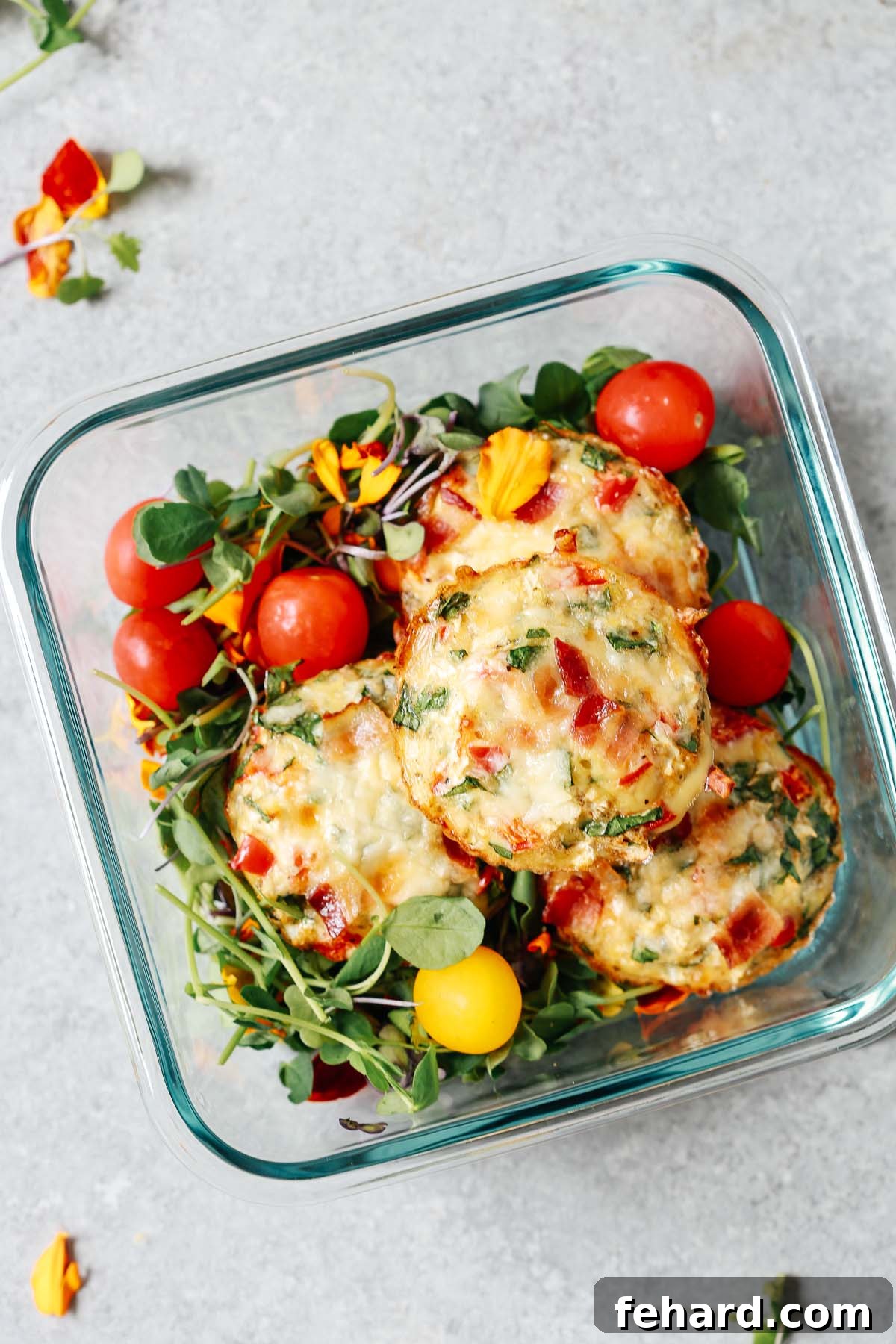 A meal prep container showcases three delicious breakfast egg muffins nestled on a bed of fresh salad greens, accompanied by bright cherry tomatoes.