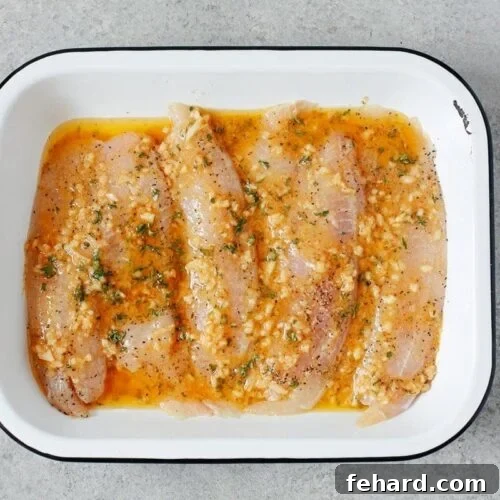 Raw tilapia inside of a white baking sheet with garlic butter sauce poured over it.