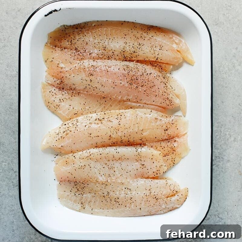 Raw tilapia fillets arranged in a single layer on a greased baking sheet, seasoned with salt and pepper.