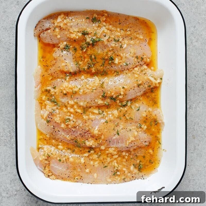 Raw tilapia fillets in a white baking dish, generously covered with garlic butter sauce before baking.