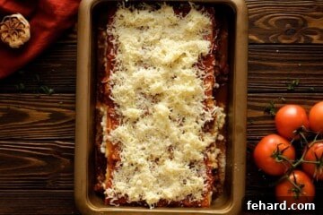 A fully layered lasagna in a 9x13 baking dish, topped with cheese and ready for baking, next to fresh tomatoes.