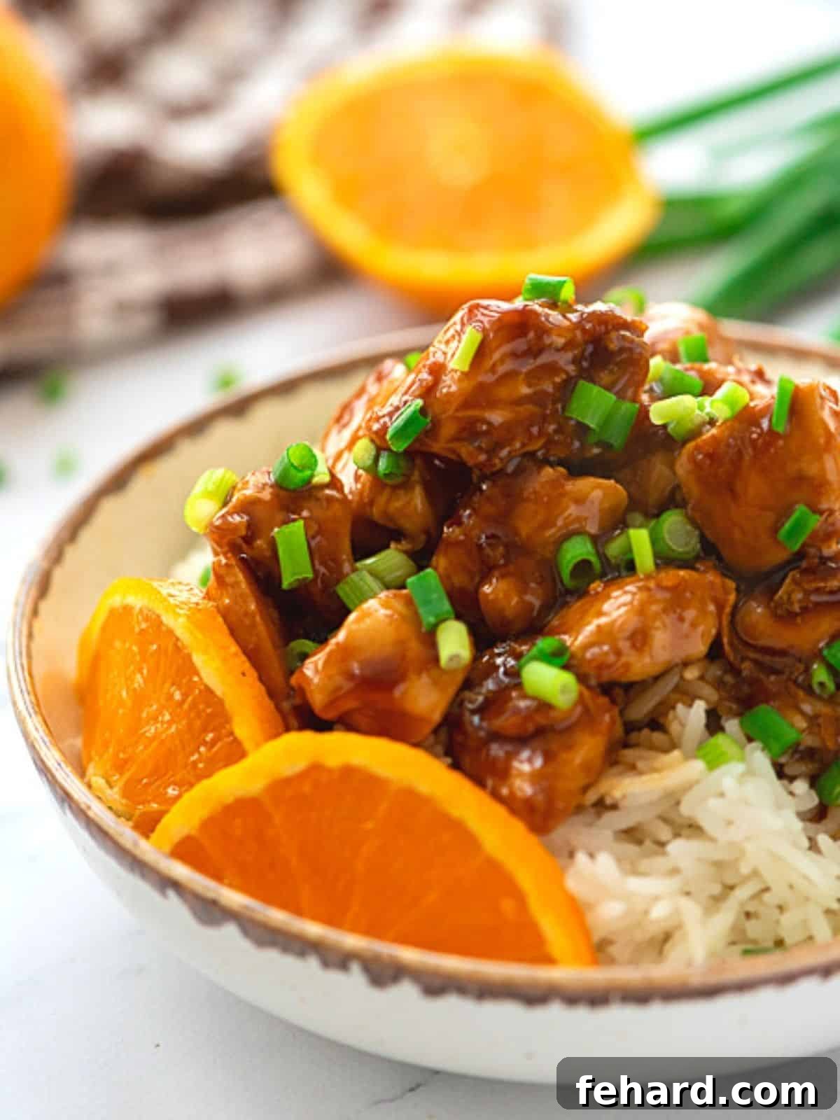 A glistening bowl of homemade orange chicken, garnished with sesame seeds and chopped green onions, served alongside fresh orange slices. The dish looks vibrant, healthy, and inviting.