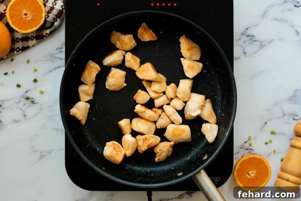 Golden-brown chicken pieces sizzling in a hot sauté pan, demonstrating the perfect sear, with fresh orange slices arranged neatly beside the pan, indicating the main flavor profile.