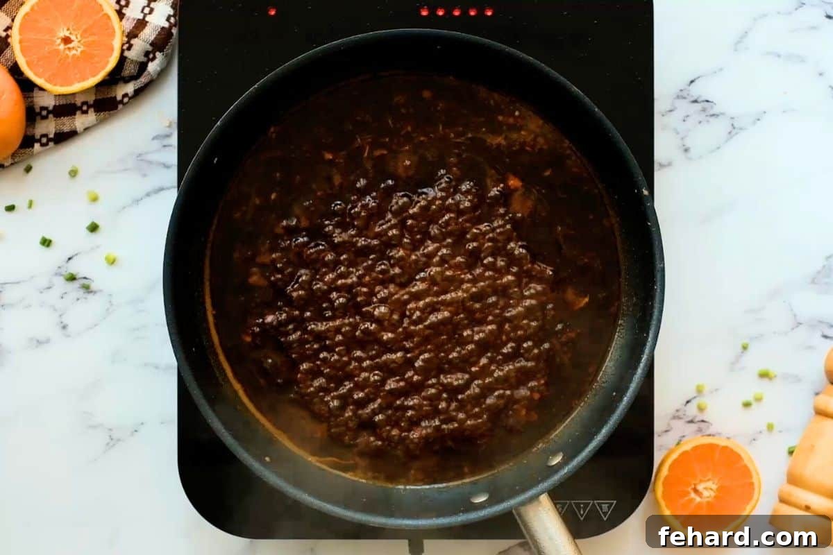 The orange chicken sauce bubbling and visibly thickening in the sauté pan, showing its transformation into a rich, glossy glaze, indicating it's ready for the chicken to be reintroduced.