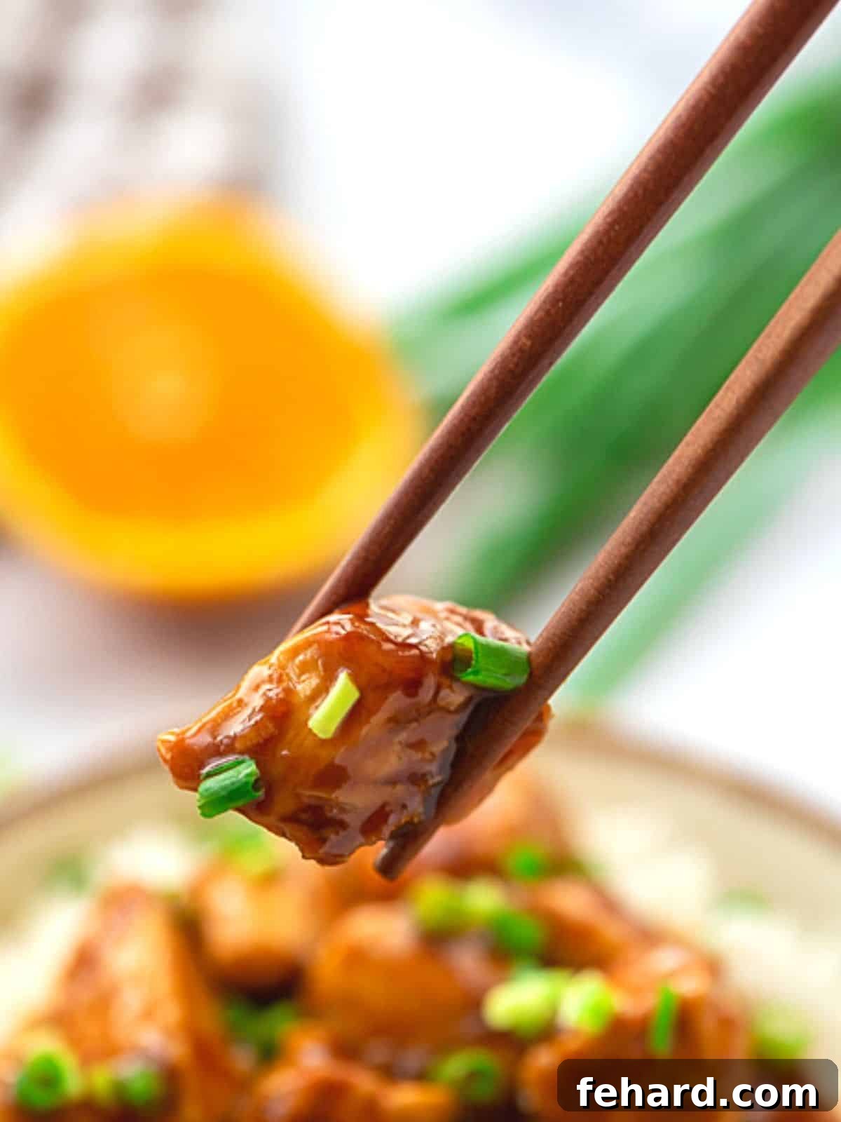 A pair of chopsticks delicately holding a piece of glistening orange chicken, showcasing its perfectly glazed texture and inviting appeal against a blurred background of a full meal.