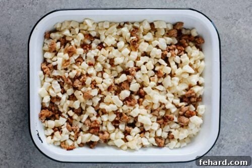 Overhead view of an assembled sausage and potato breakfast casserole in a baking dish, with layers of ingredients visible before baking.