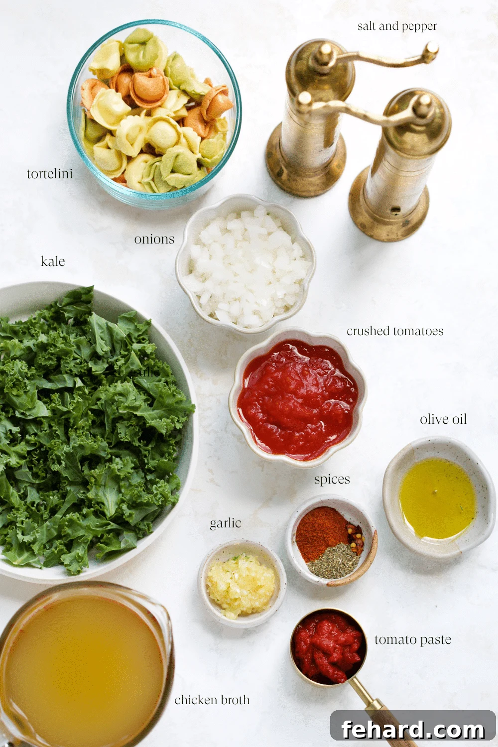 Various fresh ingredients for kale tortellini soup, including onion, garlic, tomatoes, tortellini, and kale, arranged on a wooden board.
