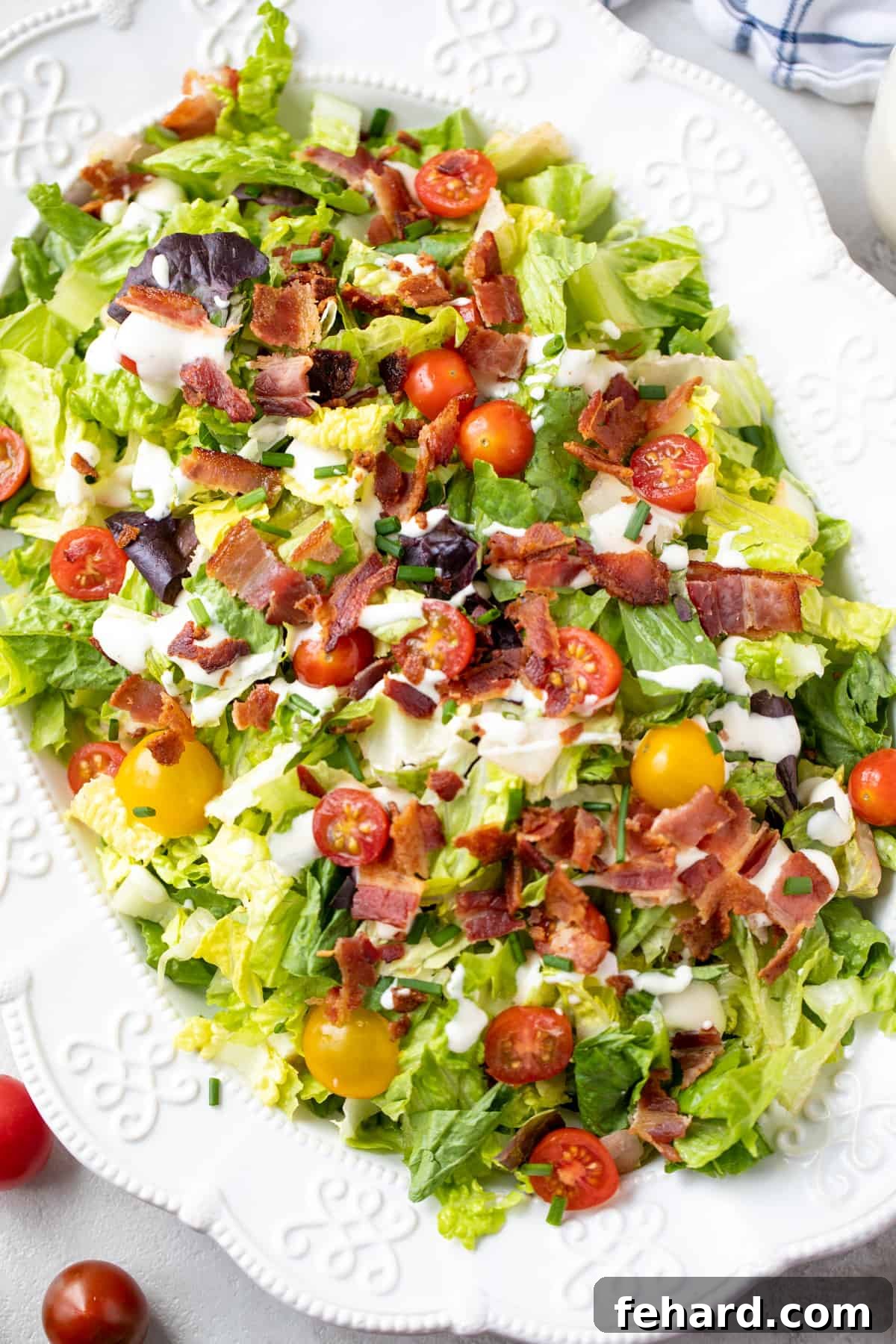 White platter showcasing a vibrant BLT Salad, garnished with fresh chives, alongside a bowl of grape tomatoes and a small jar of creamy dressing.