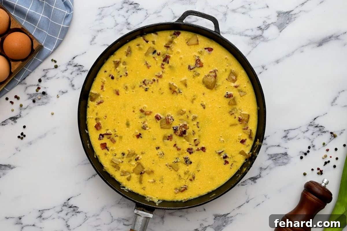 An oven-safe skillet containing the egg mixture, potatoes, crispy bacon, and grated cheese, prepared and ready for baking.