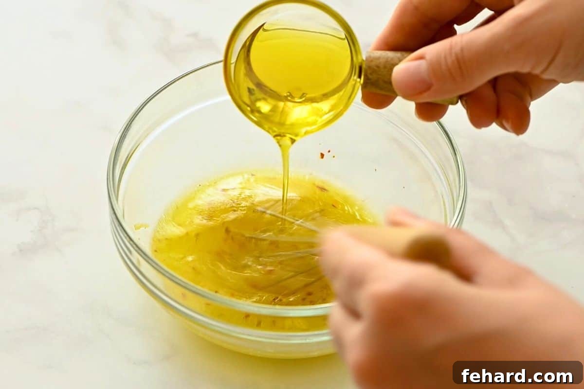 Olive oil being slowly drizzled into a bowl of lemon, garlic, and seasoning, being whisked to create an emulsified dressing for a Greek salad.