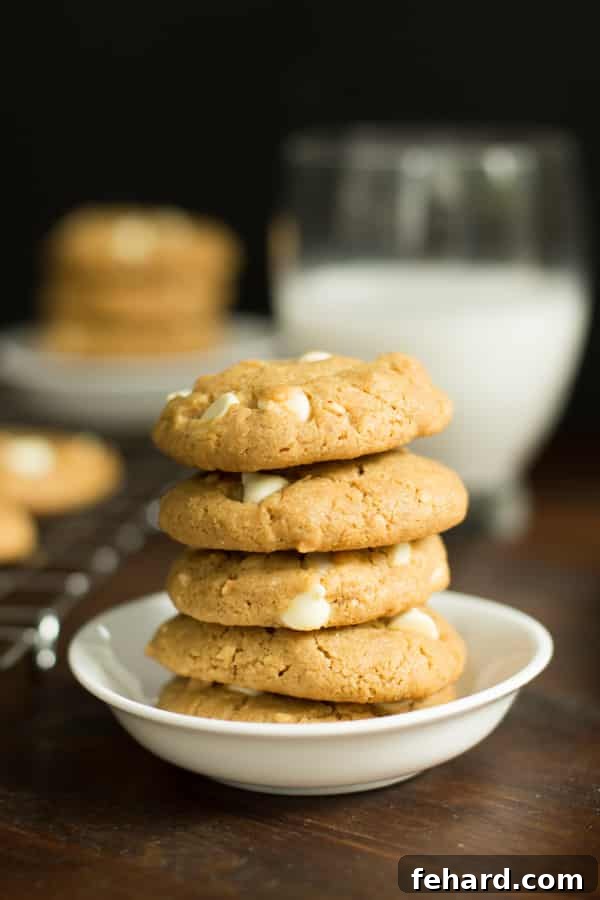 A stack of peanut butter white chocolate chip cookies in a small white bowl with a glass of milk in the background. A delightful blend of sweet and nutty flavors for your holiday spread.
