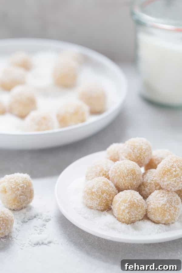Almond Coconut Snowball Cookies in a pile on a white plate with more cookies in the background. A delicate, festive, and healthier cookie option.