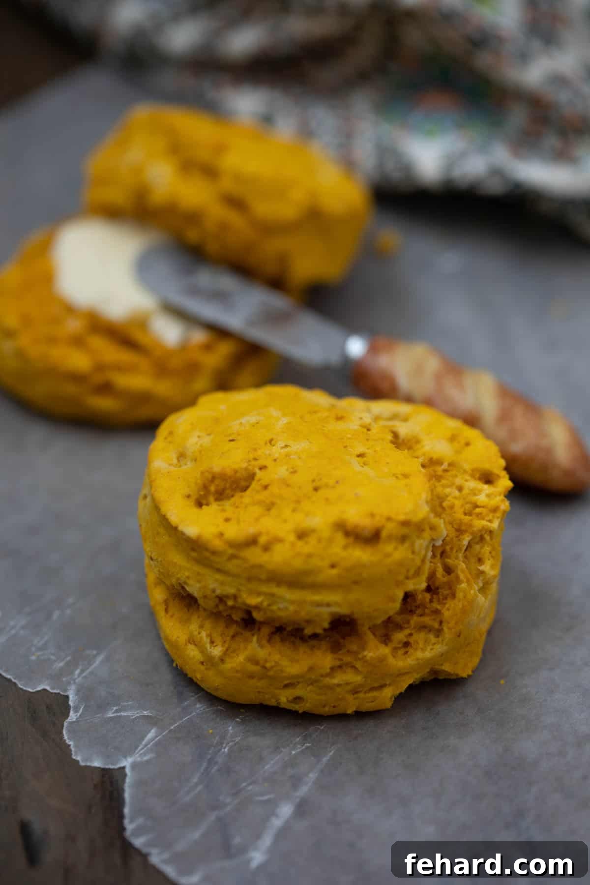 A single sweet potato biscuit resting on wax paper, with another buttered biscuit and a knife blurred in the background, highlighting its light texture.