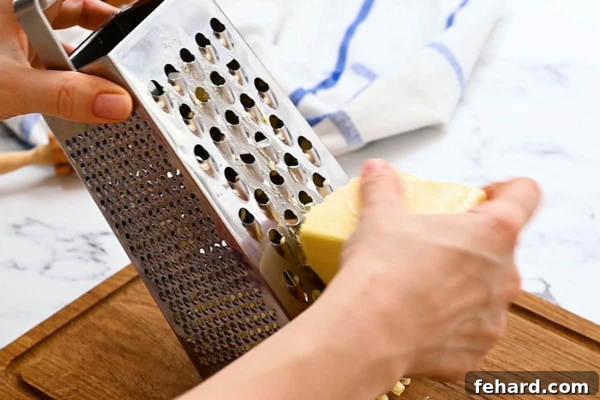 Grating cold unsalted butter with a box grater for a perfect flaky pie crust, highlighting the small, uniform pieces of butter.
