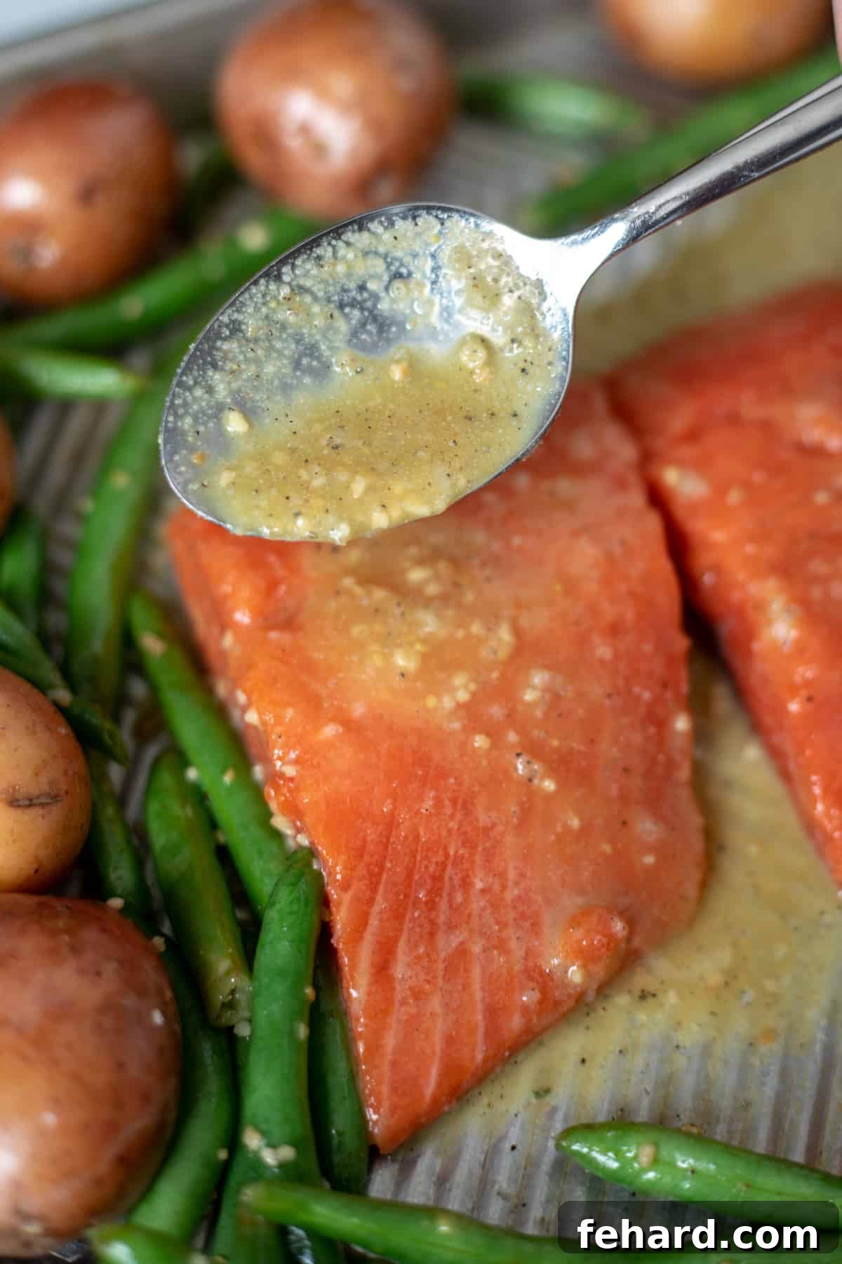 Baked Salmon with Honey Garlic Glaze A spoon drizzles the rich honey garlic glaze over a salmon fillet on a sheet pan, positioned next to roasted vegetables.