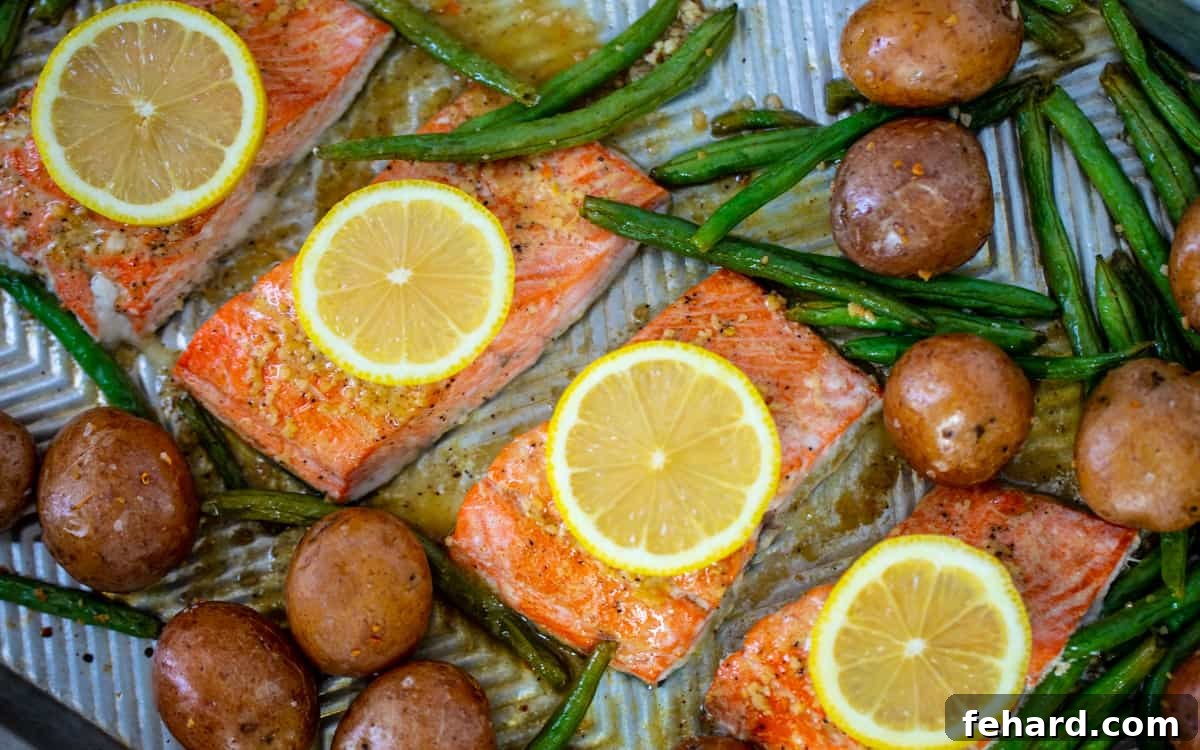 Baked Honey Garlic Salmon Four perfectly cooked salmon fillets garnished with fresh lemon slices, surrounded by roasted green beans and potatoes on a sheet pan.