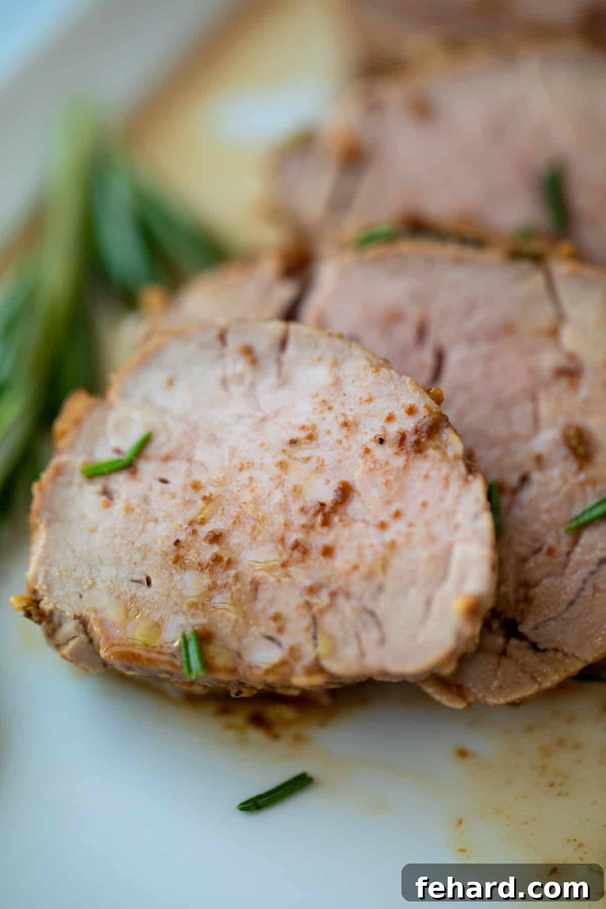 Sliced balsamic pork tenderloin on white platter with sprinkling of fresh rosemary. Each slice is perfectly cooked, showing a tender interior and a beautifully caramelized exterior, garnished with vibrant green rosemary sprigs.