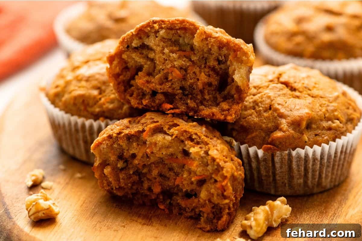 Baked Carrot Cake muffins on a cutting board with one cut open to reveal a tender, soft muffin studded with carrots and nuts.