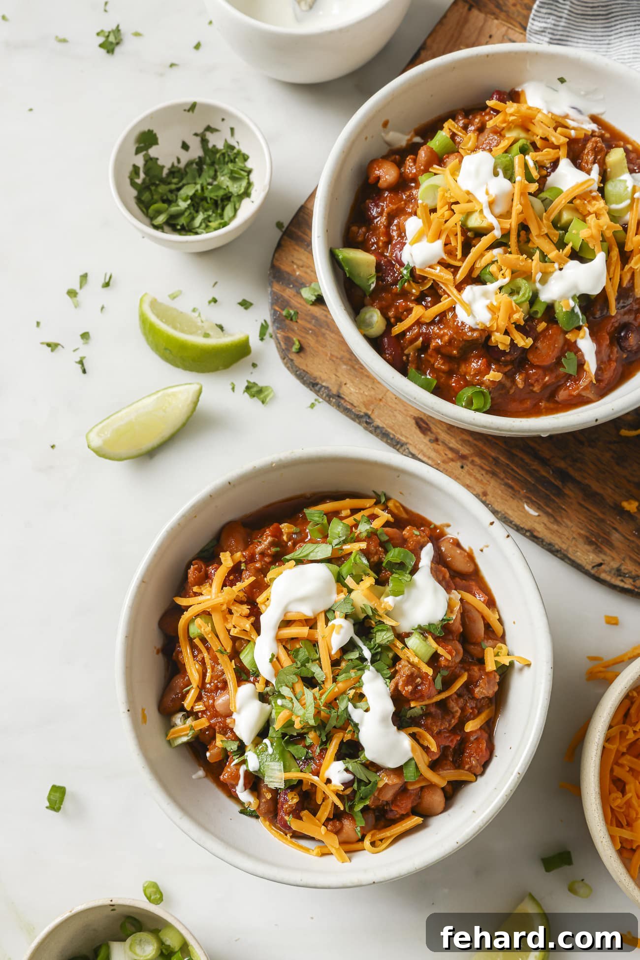 Set It and Forget It Chili 8 Two bowls of slow cooker chili, generously topped with fresh green onions, shredded cheddar cheese, and a dollop of sour cream, presented on a rustic wooden surface.