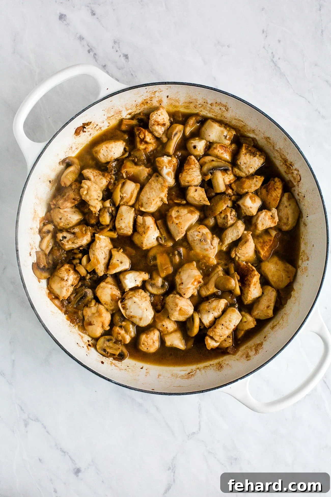 Close-up of cooked chicken, onions, broth, and mushrooms mixed in a white cast-iron braising skillet, just before the addition of sour cream.