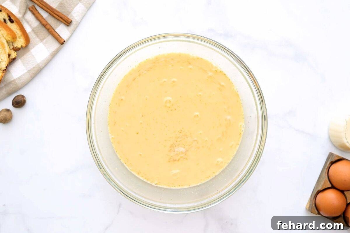 Large mixing bowl filled with whisked eggs, milk, cinnamon, nutmeg, salt, and maple syrup, creating the aromatic base for the casserole.