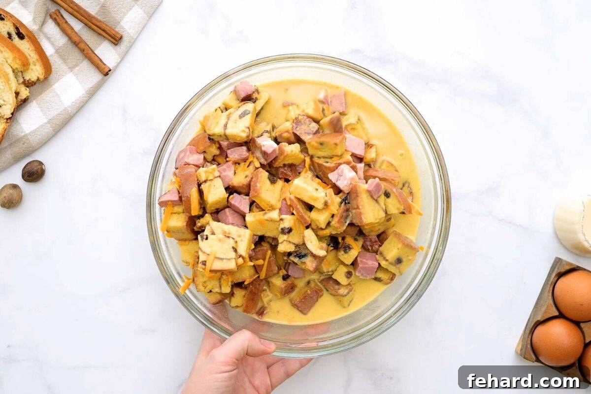 Cubed cinnamon raisin bread, diced ham, and sharp cheddar cheese being gently folded into the egg custard in a large mixing bowl, ensuring even coating.