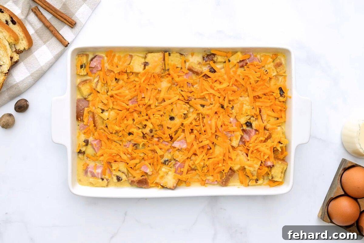 The combined mixture of cubed cinnamon raisin bread, eggs, cubed ham, and sharp cheddar cheese layered beautifully in a 9x13 inch baking dish, ready for chilling.