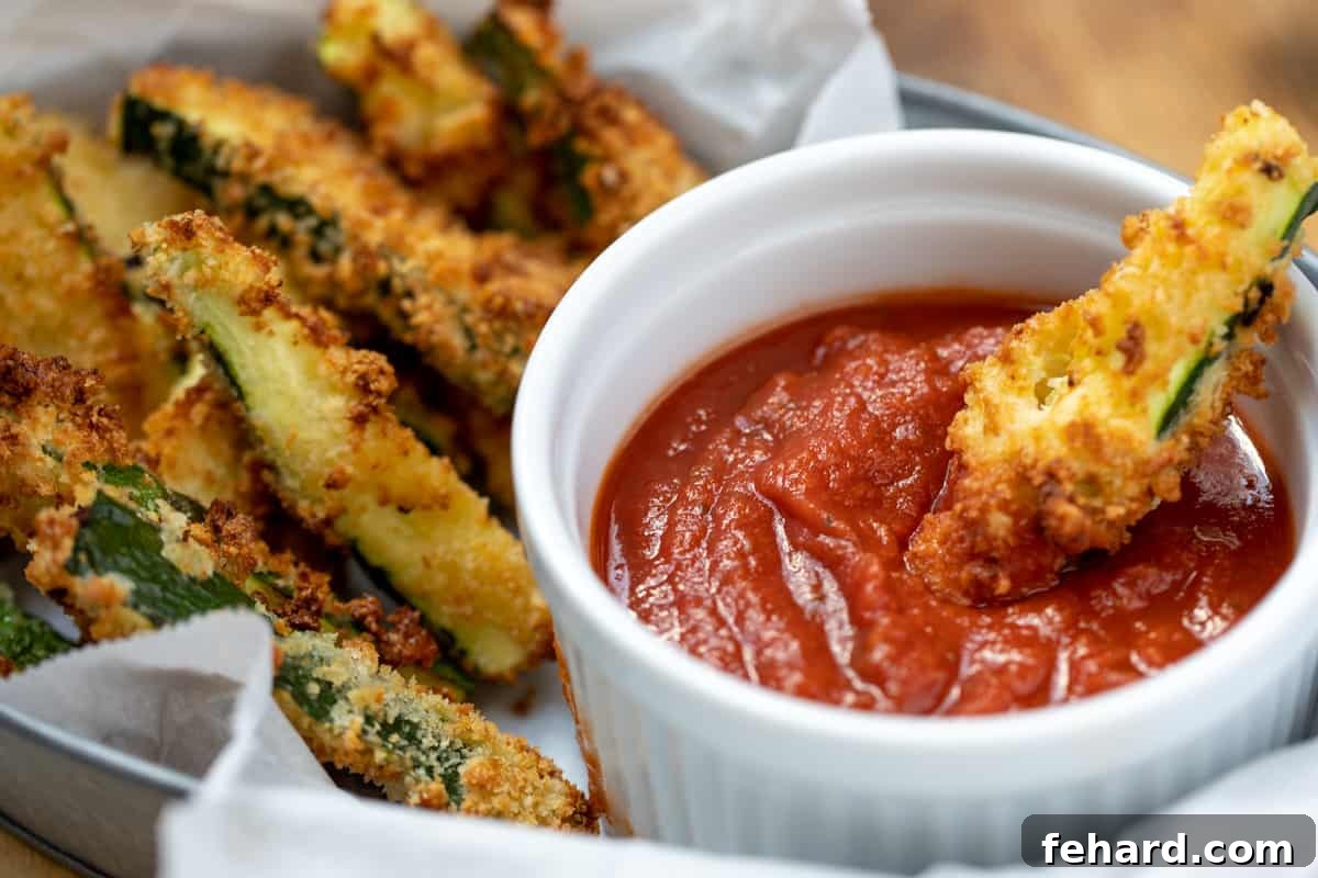 Parmesan Zucchini Fries A serving basket filled with parmesan zucchini fries, with one fry being dipped into a small bowl of marinara sauce.