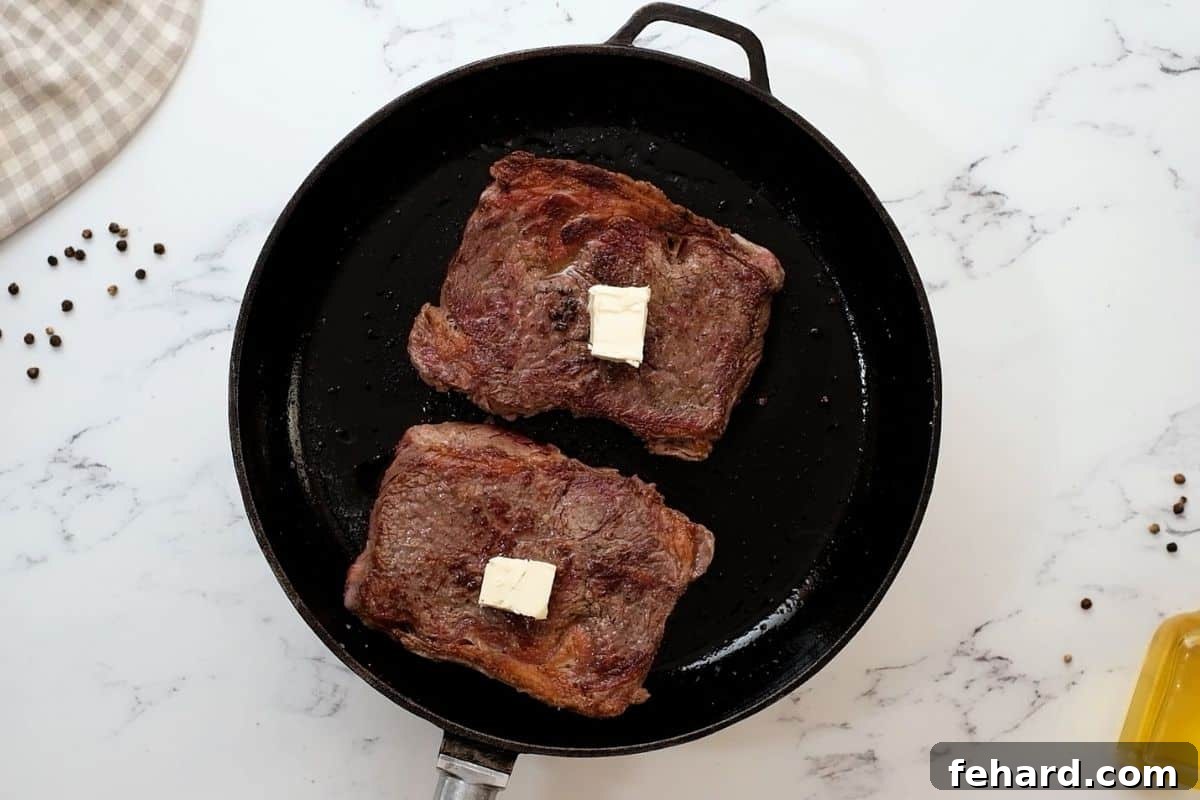 Two beautifully seared ribeye steaks, glistening with melted butter, resting in a hot cast-iron skillet after being flipped, showcasing their perfect golden-brown crust.