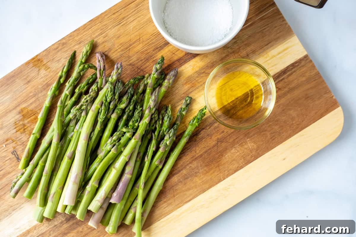 Freshly trimmed asparagus stalks arranged on a wooden cutting board, next to a bottle of extra virgin olive oil and a small bowl of kosher salt.